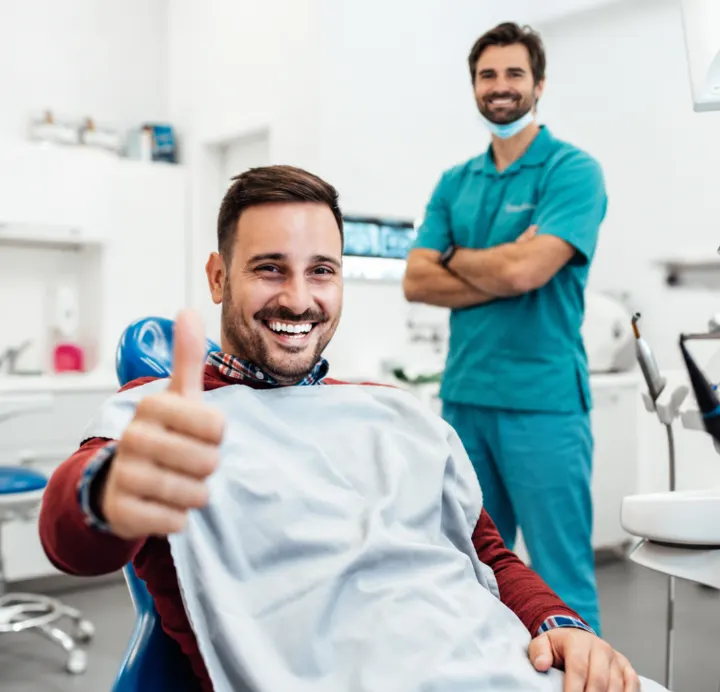 Smiling man in a dental chair giving a thumbs up with his dentist behind him